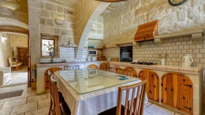 A kitchen with stone walls, wooden cabinets, a glass-top table, and a clock on the wall. - Ta'leli Pool Farmhouse in Gozo (Gharb)