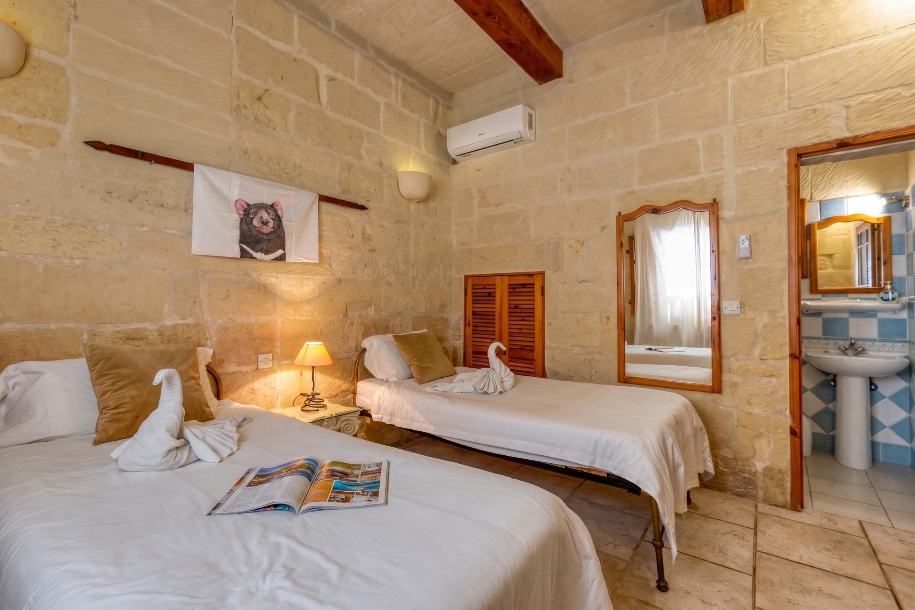 A room with two beds, a wall-mounted air conditioner, a wooden door, a mirror, and a bathroom with a sink and tiled walls.