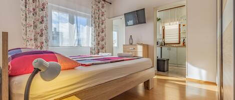 A bedroom with a bed, a desk with a lamp, a dresser, and a TV mounted on the wall.