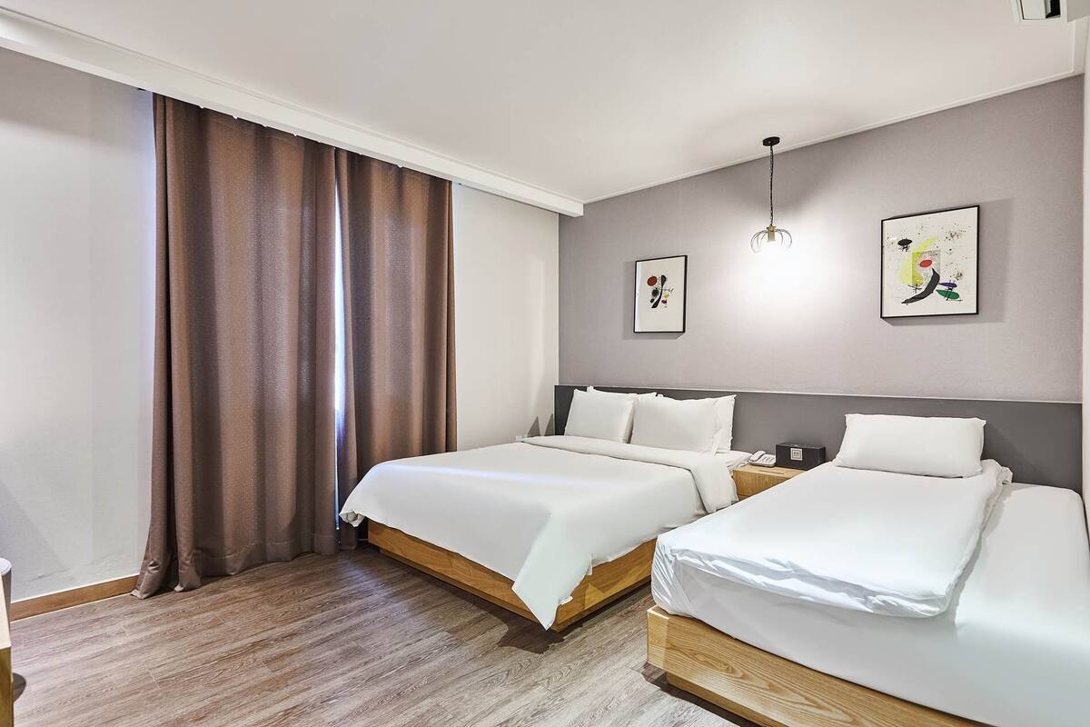 A hotel room with two beds, wooden flooring, a large window with curtains, and two wall-mounted artworks.