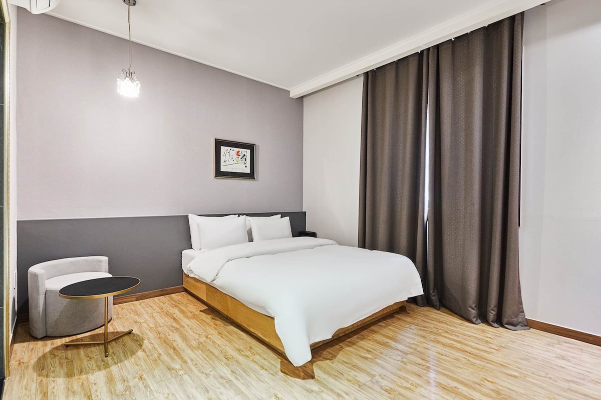A modern hotel room with a wooden floor, a bed with white linens, a small round table, a chair, a framed picture on the wall, and a large window with curtains.