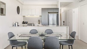 Uma cozinha moderna com uma ilha central, eletrodomésticos de aço inoxidável e uma área de jantar com mesa de vidro e cadeiras cinzas.