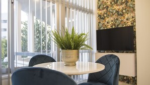 A room with a round table, two blue chairs, a potted fern, a television on the wall, and a balcony with vertical blinds. - STAY Glam Skin Studio (Nicosia)