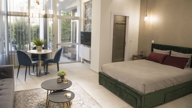 A modern bedroom with a large bed, a dining area with chairs, a small table with a plant, and a view through large windows. - STAY Glam Skin Studio (Nicosia)