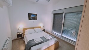 A bedroom with a bed, bedside lamps, a window with blinds, a radiator, and a painting on the wall. - STAY Mystique City Penthouse (Limassol)
