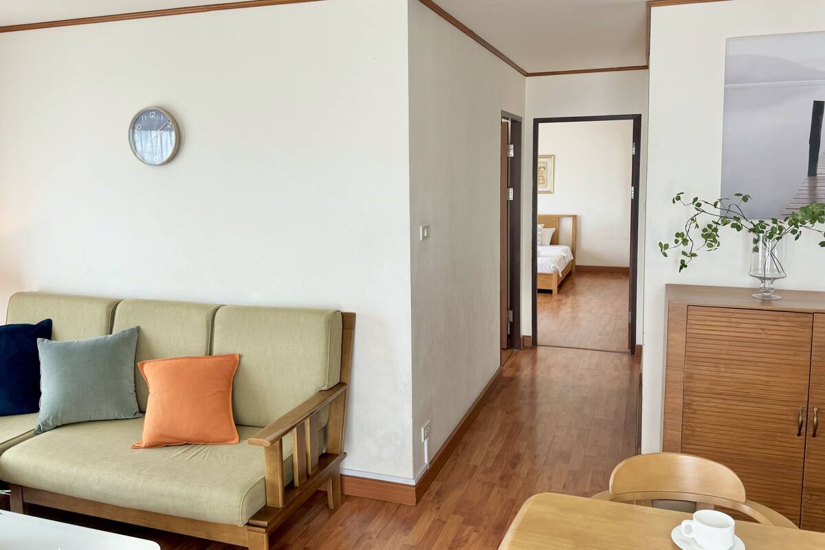 Una habitación con sofá, una mesa de comedor con una taza, un armario de madera y una cama al fondo.