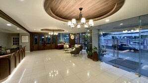 A modern hotel lobby with a wooden ceiling, a chandelier, a reception desk, seating area, and a glass wall offering a view of the street. - Sriracha Avenue (Si Racha)