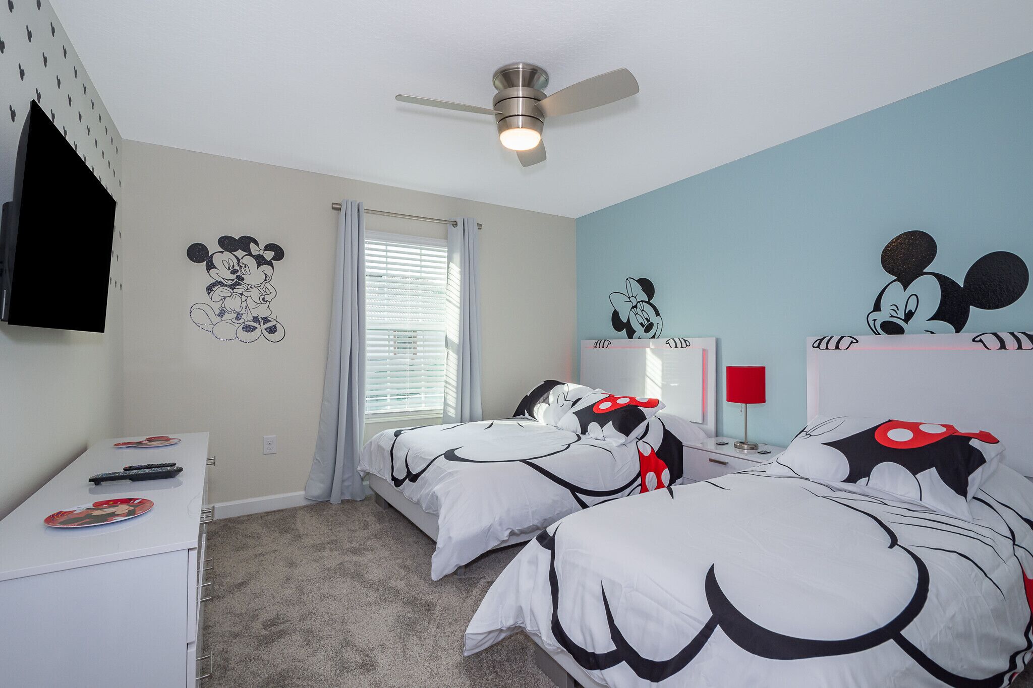A bedroom with two beds, a ceiling fan, a TV mounted on the wall, and Mickey Mouse wall decals.