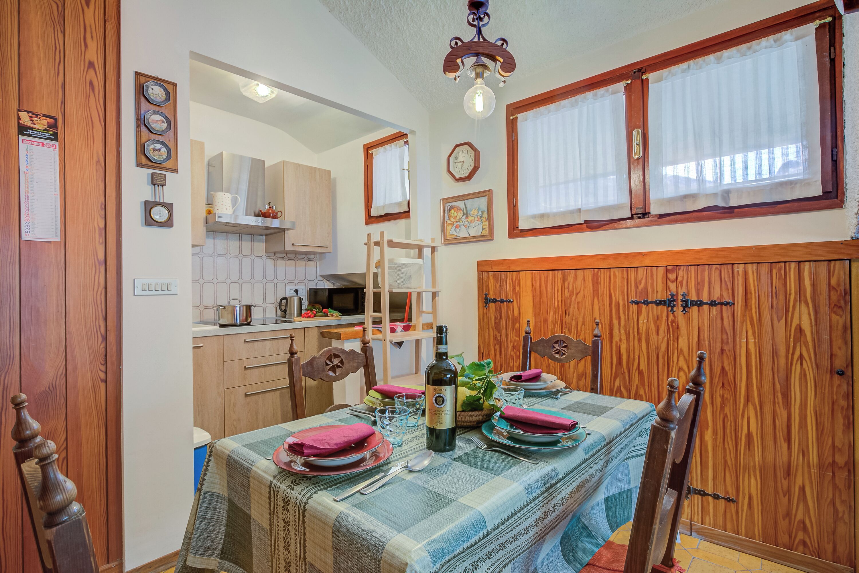 A dining area with a table set for four, wooden chairs, a chandelier, and a kitchen in the background.