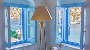 A room with a sofa, a lamp, and a small table with decorative items. There are two windows with blue shutters, one of which is open. - Seafront - Harbour view Nikolakis (Symi)