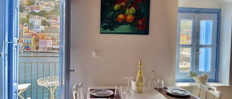 A dining area with a view of colorful buildings through an open door, a table set with plates and glasses, and a painting of fruit on the wall.