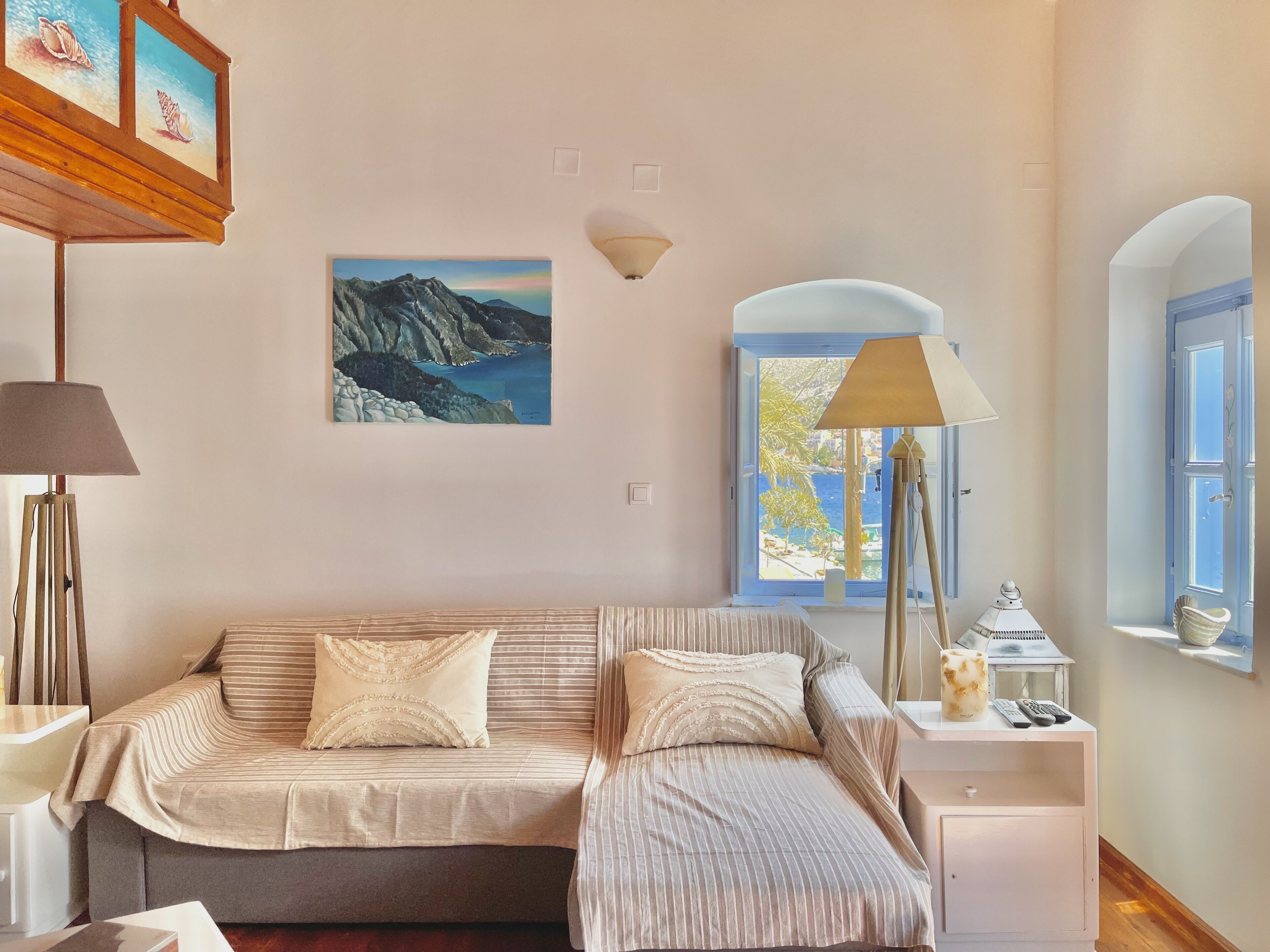 A bedroom with a bed, a nightstand, a lamp, a picture of mountains, and a window with a view of the sea.