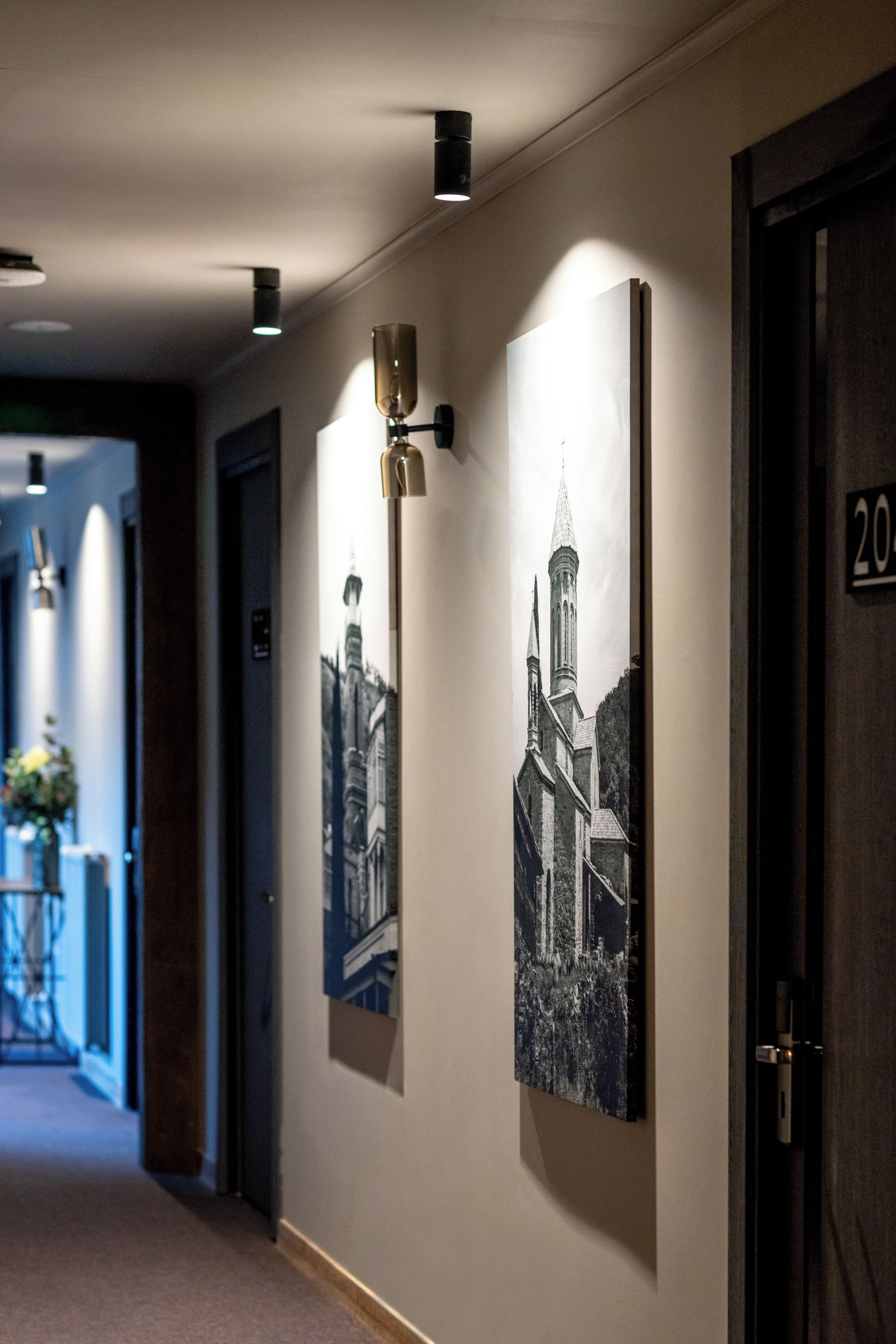 Een gang met zwarte-en-witte foto's in lijsten aan de muur, een deur met de aanduiding '20' en een lamp erboven.