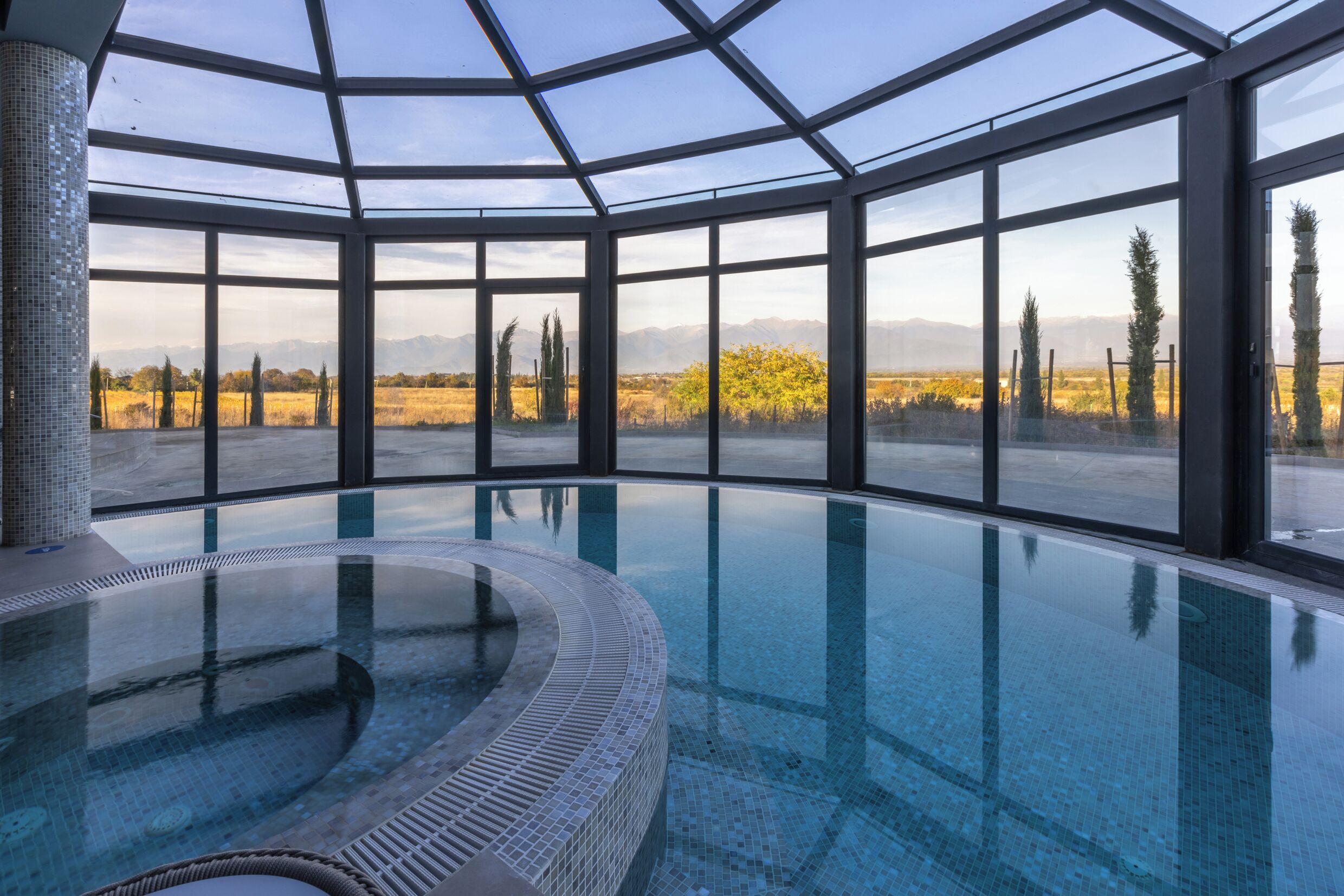 A large indoor swimming pool with a glass ceiling, surrounded by large windows offering a view of a natural landscape.