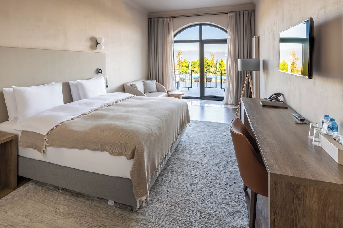Une chambre d’hôtel avec un grand lit, un bureau, une chaise, une télévision et une vue sur une piscine et des arbres.