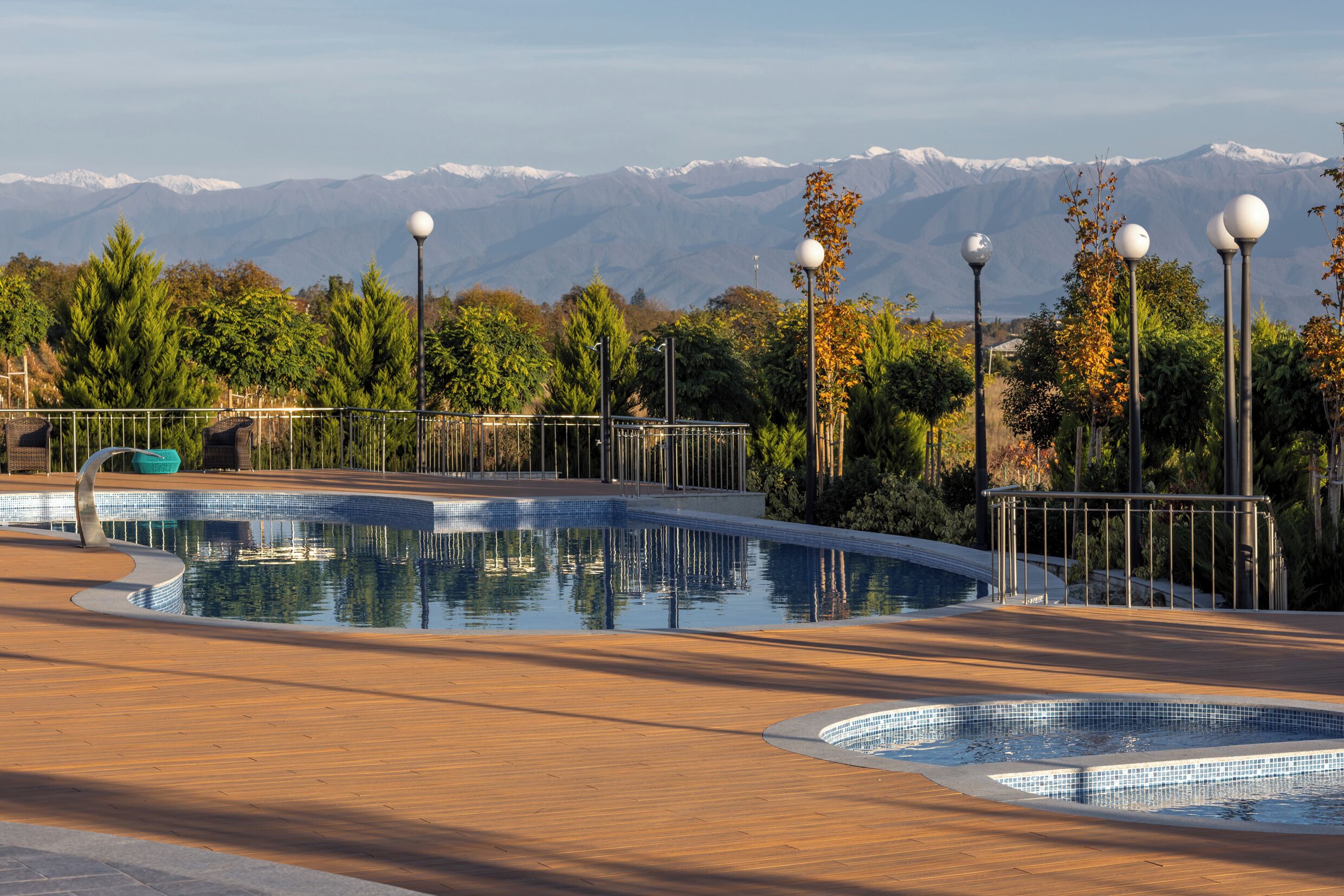 A swimming pool with a wooden deck, surrounded by trees and street lamps, with a mountain range in the background.