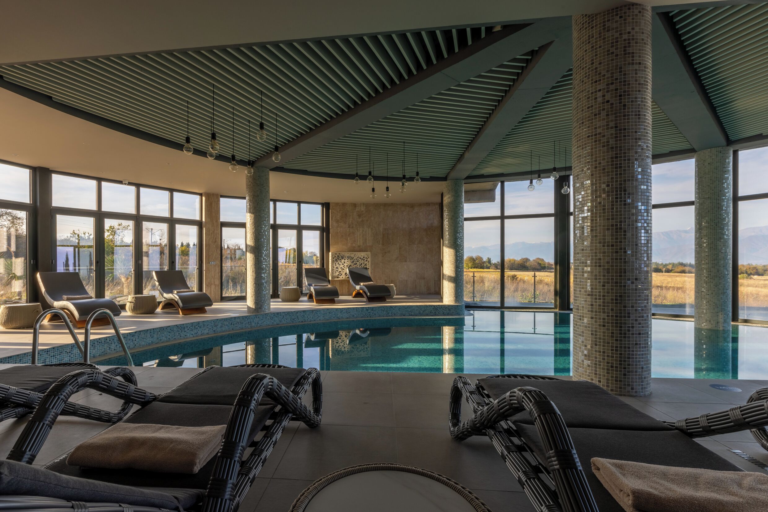 A modern indoor swimming pool with lounge chairs and large windows offering a view of the landscape.