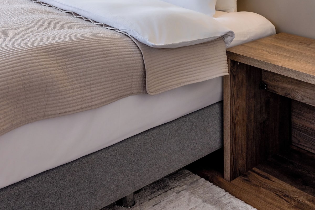 Un lit bien fait, avec du linge de lit blanc, une table de chevet en bois et des lampes fixées au mur.