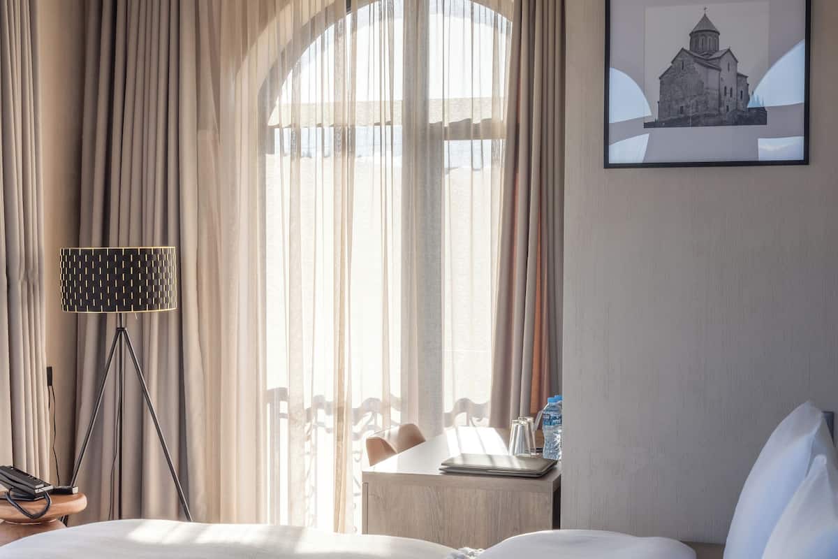 Une chambre d’hôtel avec un grand lit, un bureau avec une lampe, une chaise, une petite table avec une bouteille d’eau et un tableau encadré au mur.