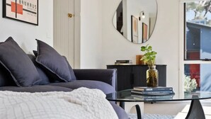 A modern living room with a dark grey sofa, a glass coffee table, and a white knitted throw. A framed artwork and a mirror are on the wall, and a vase with green plants is on the table. - Boxy's Beach House: Perfect Location - Sea ViewsPeace & Quiet - Walk Anywhere (Lorne)