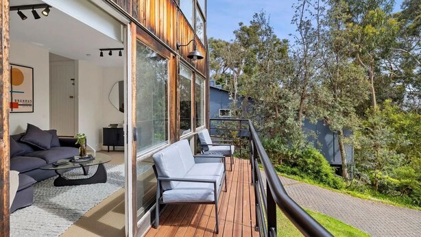 A modern outdoor deck with a sofa and chairs, surrounded by glass walls and wooden deck flooring. - Boxy's Beach House: Perfect Location - Sea ViewsPeace & Quiet - Walk Anywhere (Lorne)