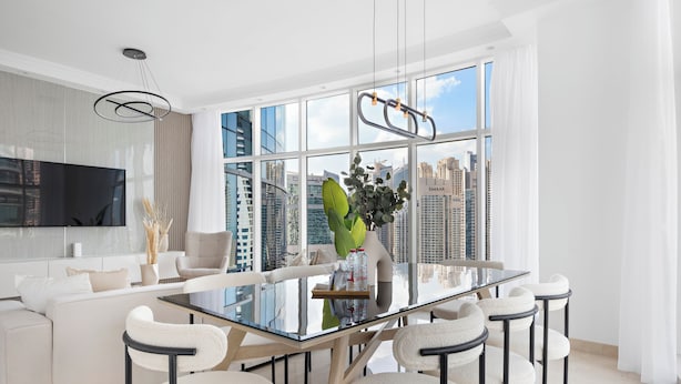 A modern dining area with a glass table, white chairs, and a city view through large windows. - Silkhaus Trident Oceanic - Dubai Marina (Dubai)