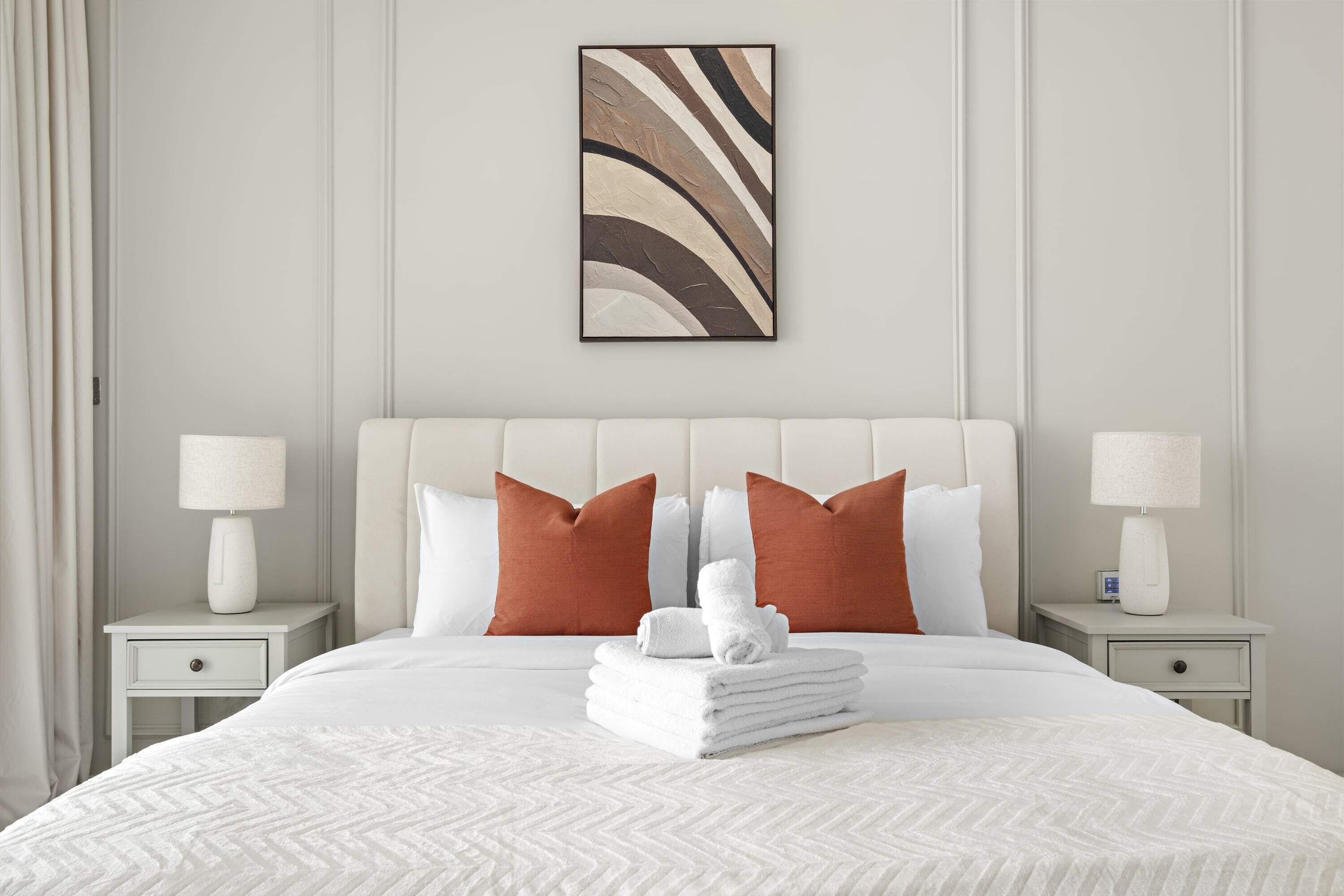 Ein ordentlich bezogenes Bett mit weißer Bettwäsche und zwei Kissen, ein an der Wand befestigtes abstraktes Kunstwerk und zwei Nachttische mit Lampen.
