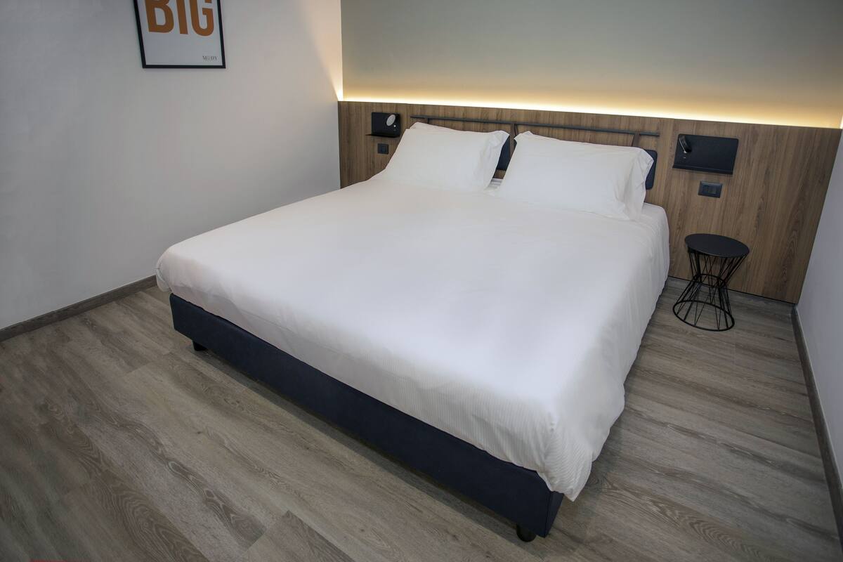 A modern hotel room with a large bed, wooden headboard, a small side table, and a framed picture on the wall.