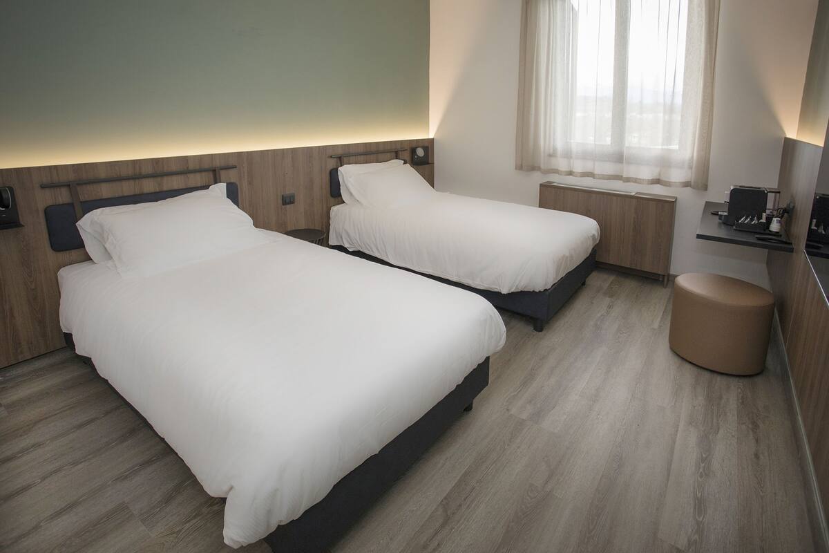 A hotel room with two beds, a small table, a round ottoman, a window with sheer curtains, and a wall-mounted light.