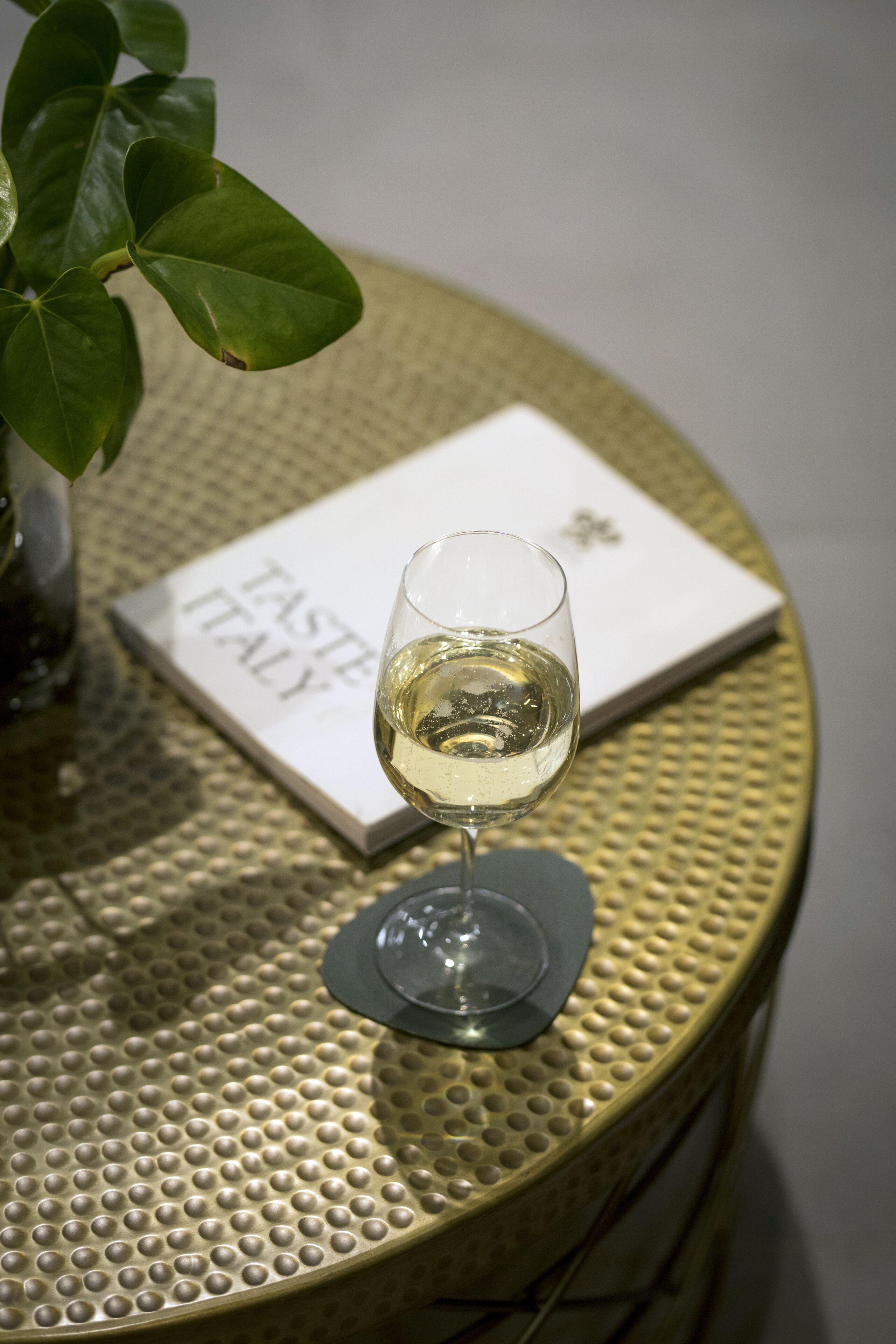 A glass of white wine on a textured round table with a booklet titled "TASTE ITALY" beside it.