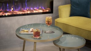 Una mesa de vidrio con un sándwich y una bebida, un sofá amarillo y una chimenea con llamas.