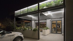 Un edificio moderno con fachada de vidrio, un automóvil estacionado al frente y un área de entrada bien iluminada.