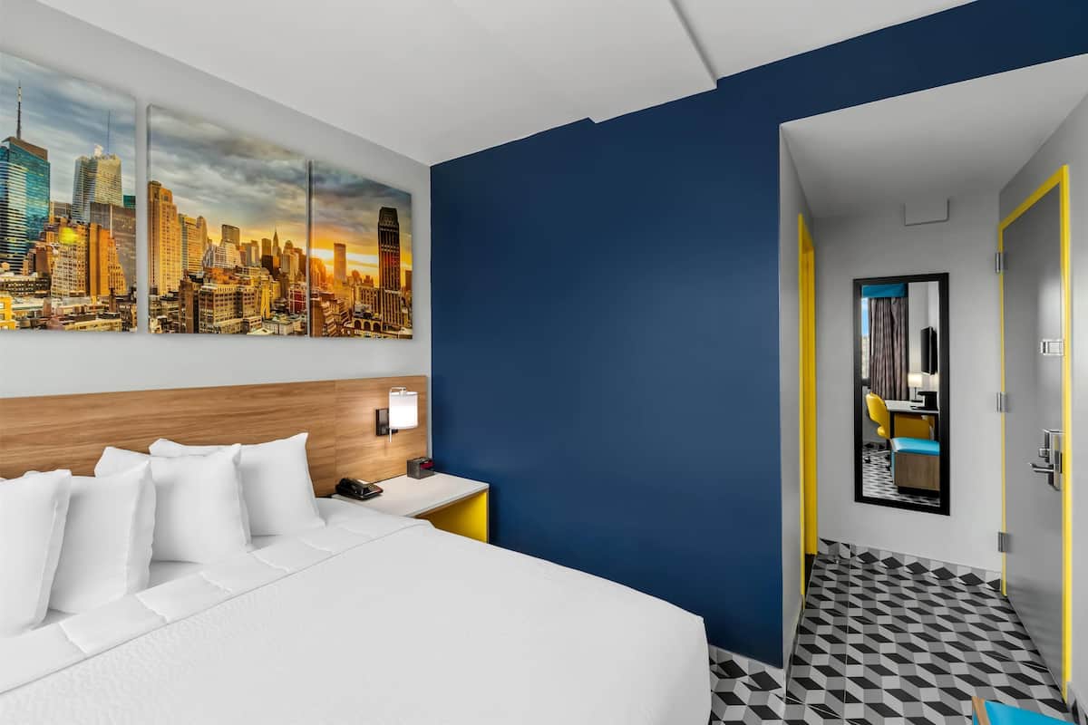 A hotel room with a large bed, a cityscape painting, a wooden headboard, a nightstand, and a bathroom with a checkered floor.
