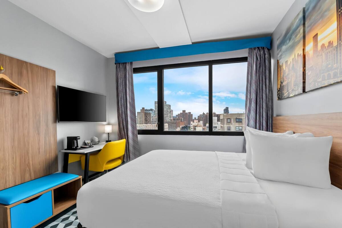 A hotel room with a large bed, a desk with a chair, a television, and a view of the city skyline.