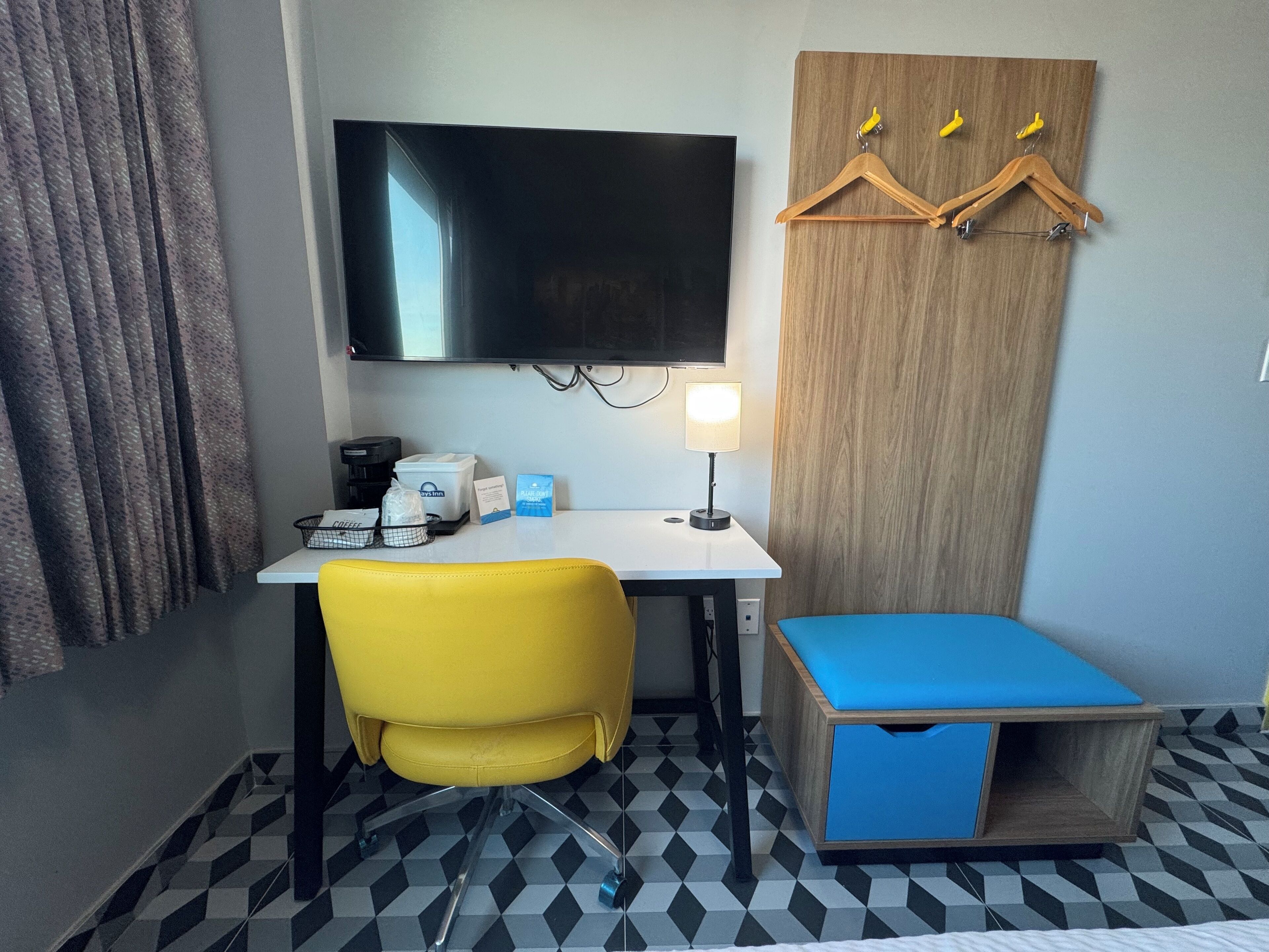 A compact hotel room with a flat-screen TV, a small desk with a coffee maker, a yellow chair, a blue bed, and a wooden wardrobe with hangers.