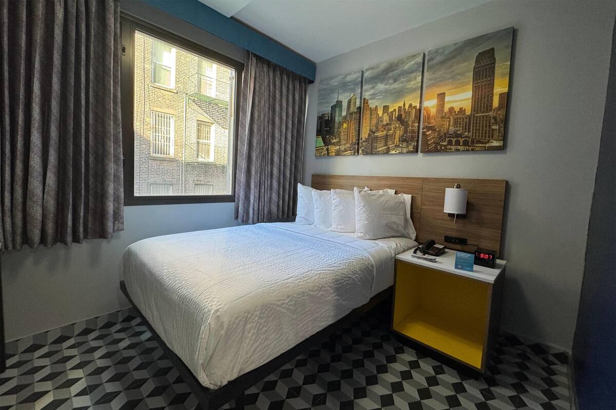 A hotel room with a bed, a nightstand, a window with curtains, and a cityscape painting on the wall.