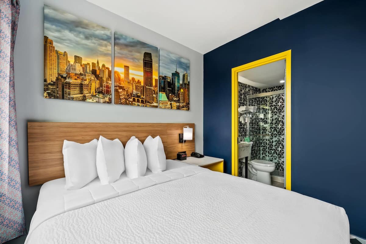 A hotel room with a large bed, a cityscape painting above the bed, a wooden headboard, a bedside table with a lamp, and a bathroom with a yellow frame.
