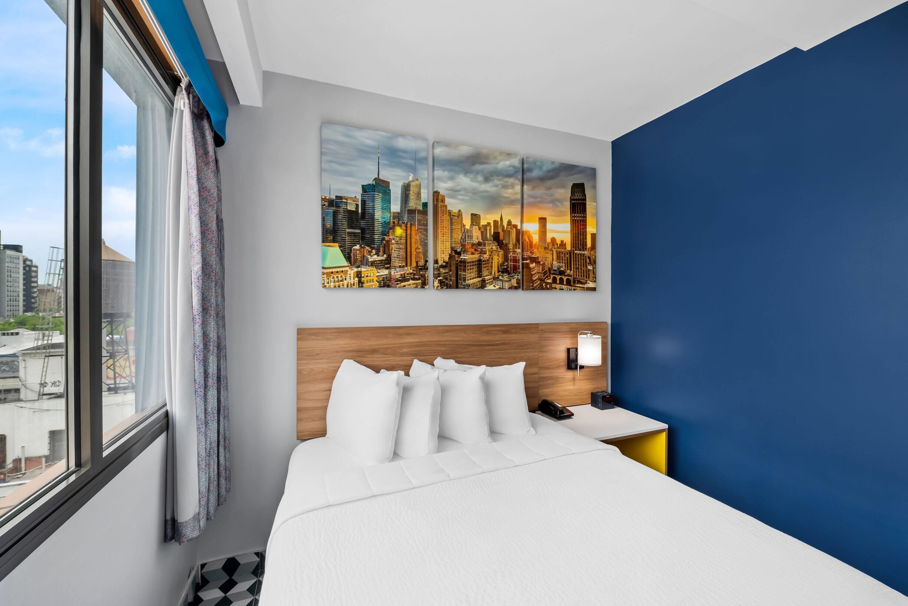 A hotel room with a large bed, a cityscape painting on the wall, a wooden headboard, and a nightstand with a phone.