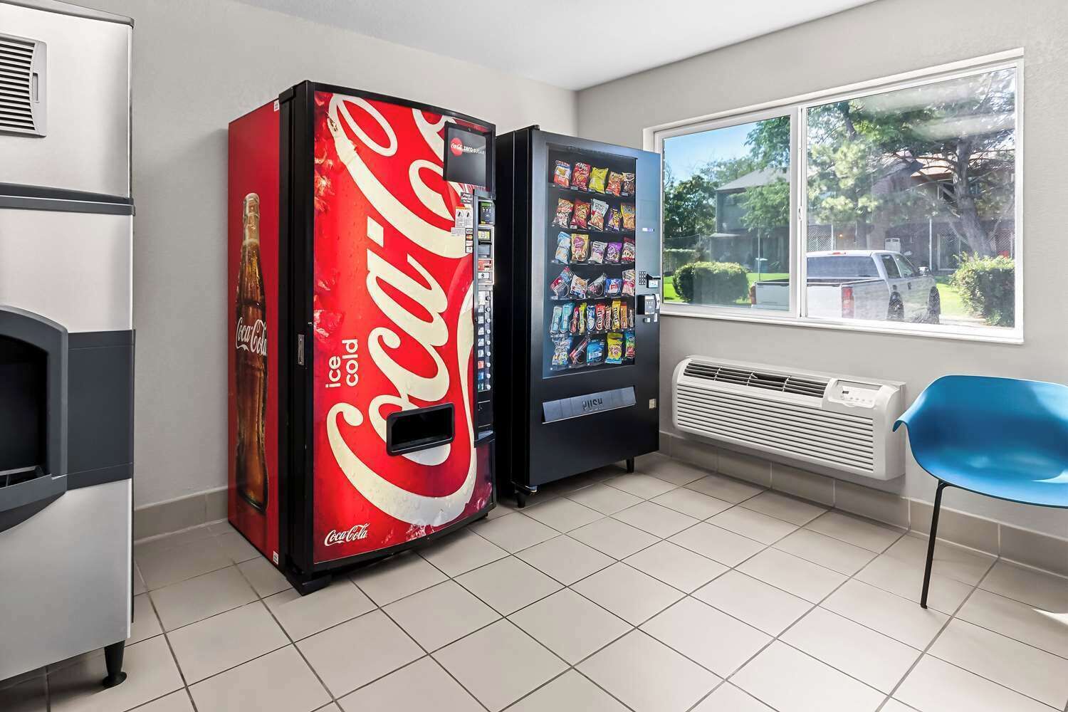 A room with a large Coca-Cola vending machine, a smaller snack vending machine, a blue chair, and a window with a view of the outside.