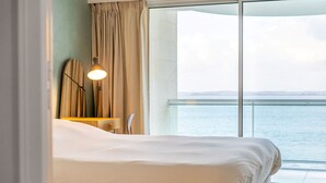 A hotel room with a bed, a lamp, a surfboard, and a view of the sea.