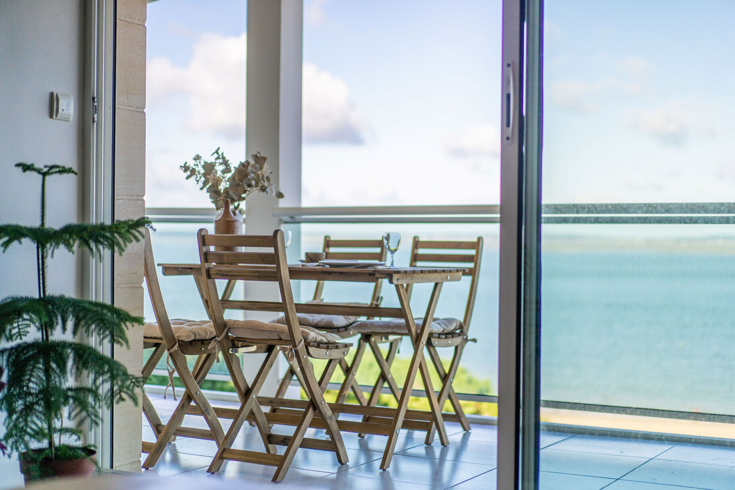 A balcony with a wooden table and chairs, a potted plant, and a view of the sea.