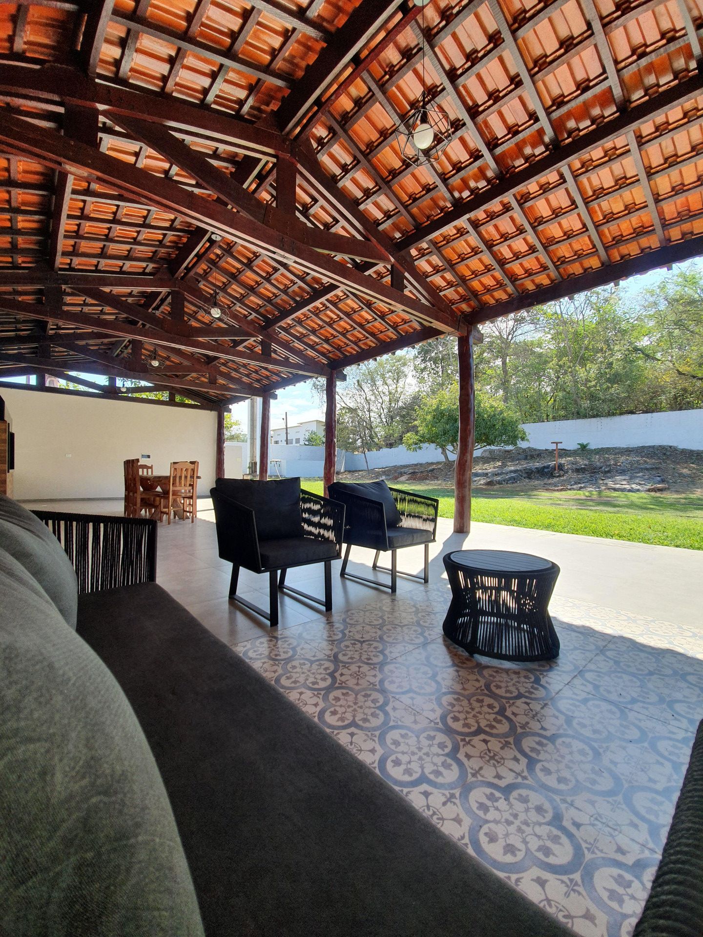 An open-air covered patio with a patterned floor, two black chairs, a round table, and a view of greenery outside.