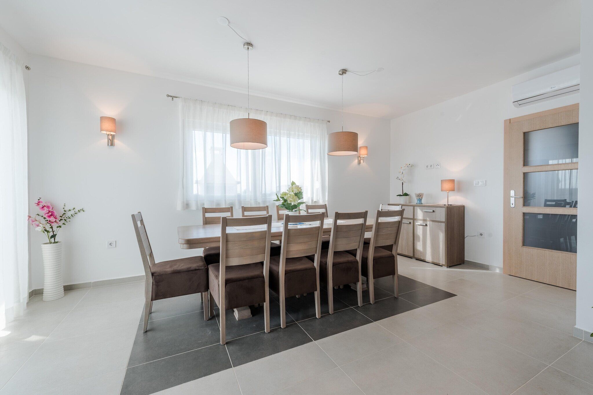 A modern dining area with a rectangular table, six chairs, pendant lighting, a vase with flowers, and a kitchen area with a wooden cabinet and a window with sheer curtains.