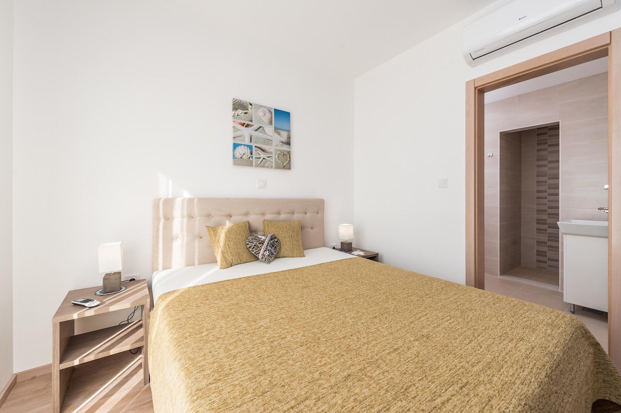 Een keurig opgemaakt slaapkamer met een bed, nachtkastjes, een kunstwerk aan de muur en een badkamer zichtbaar door een open deur.