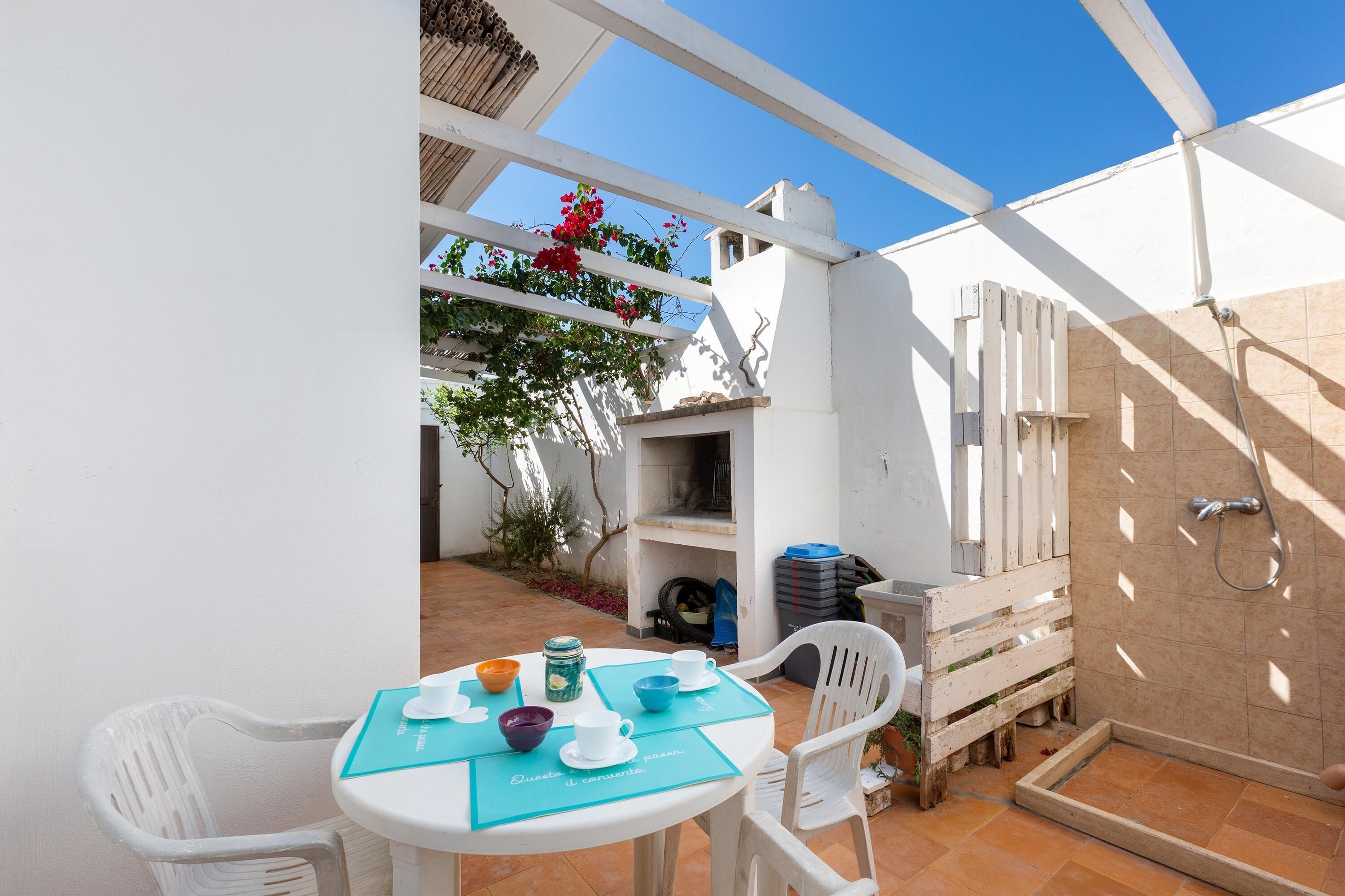 Un patio con una mesa lista para dos, una silla blanca y un pequeño jardín con cerca de madera y flores rojas.