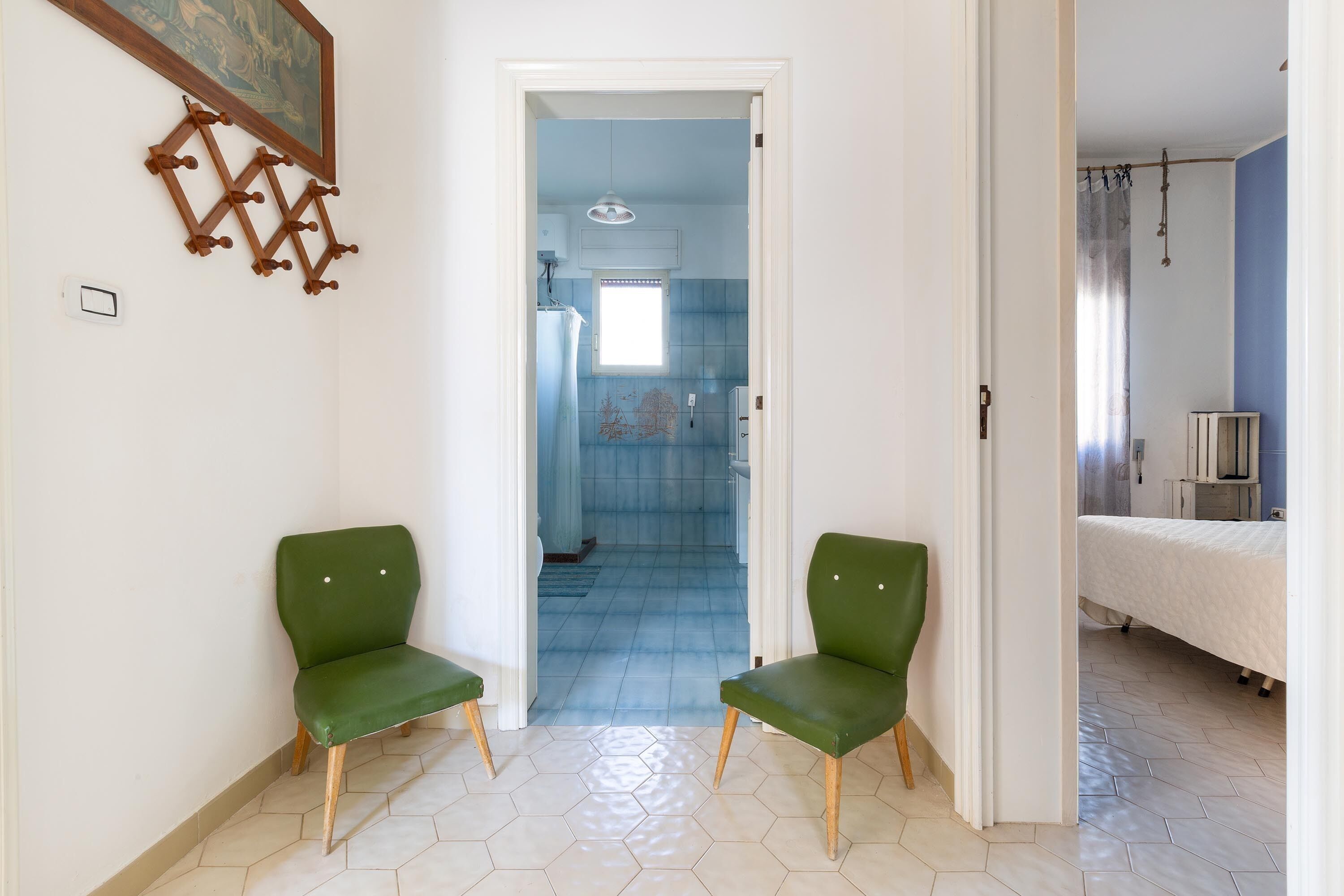 Een gang met twee groene stoelen, een deur naar een badkamer en een bed in de aangrenzende kamer.