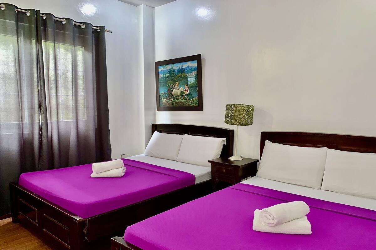 A hotel room with two beds, each with a purple bedspread and white pillows, a painting on the wall, and a nightstand with a lamp.