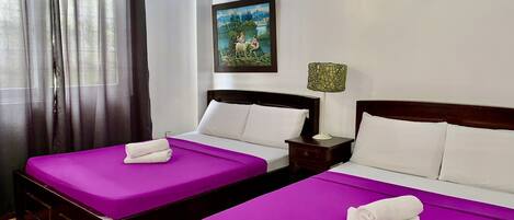 A hotel room with two beds, each with a purple bedspread and white pillows, a painting on the wall, and a nightstand with a lamp.