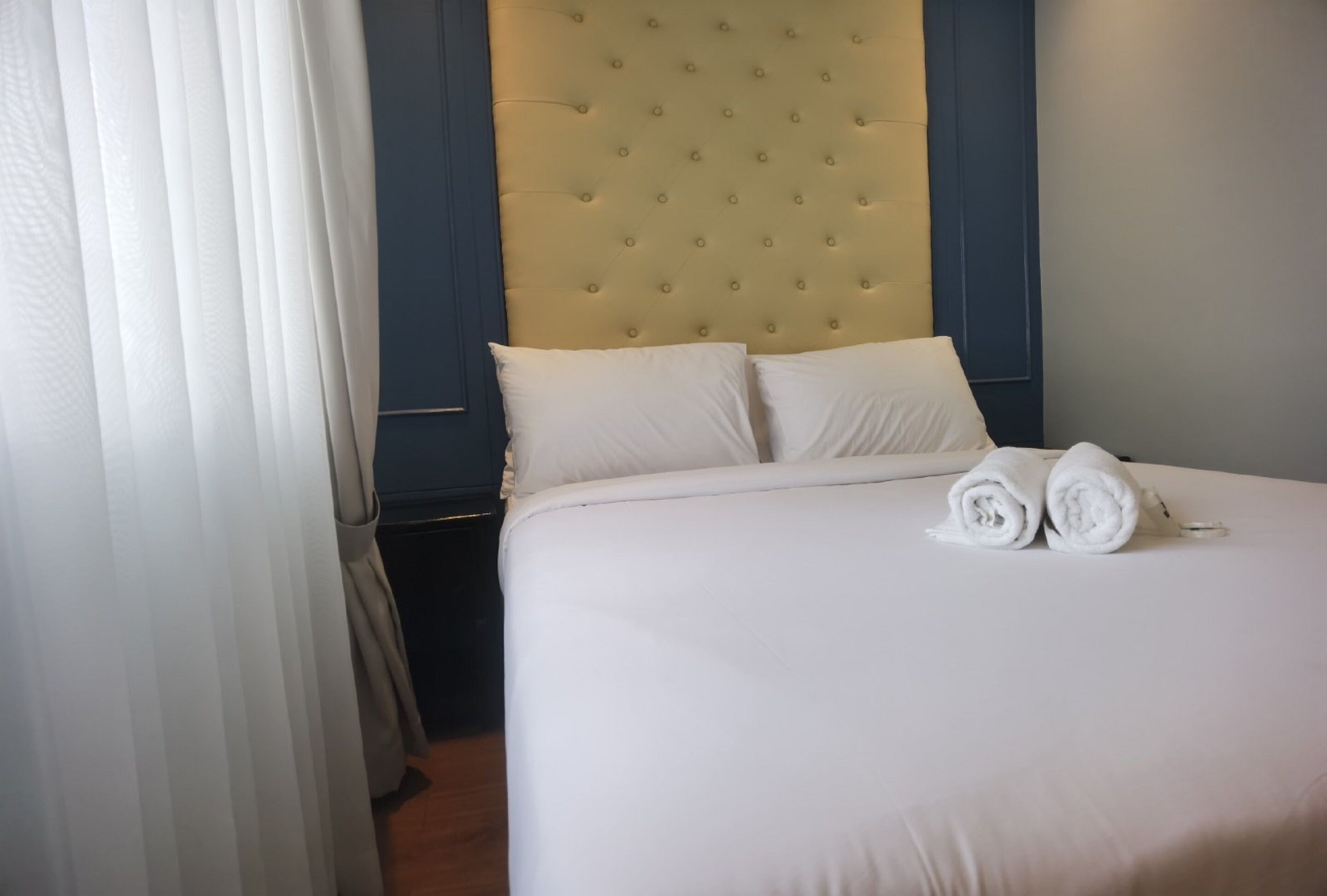 A neatly made bed with white linens and two rolled towels, a tufted headboard, and a dark blue wall.