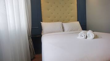 A neatly made bed with white linens and two rolled towels, a tufted headboard, and a dark blue wall.